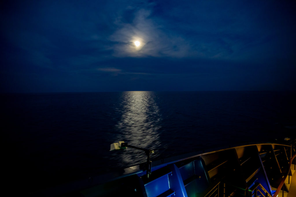 Blick auf das Meer bei Nacht von Bord der Mein Schiff 3 von TUI Cruises. Im Hintergrund ist der Vollmond zu sehen, sein Licht spiegelt sich auf dem fast schwarzen Wasser.