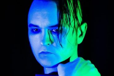 Ein Portrait von System Syns Clint Carney. Er hält etwas in seinen Armen vor der Brust verschränkt, das Gesicht ist komplett in grünes und blaues Licht getaucht.