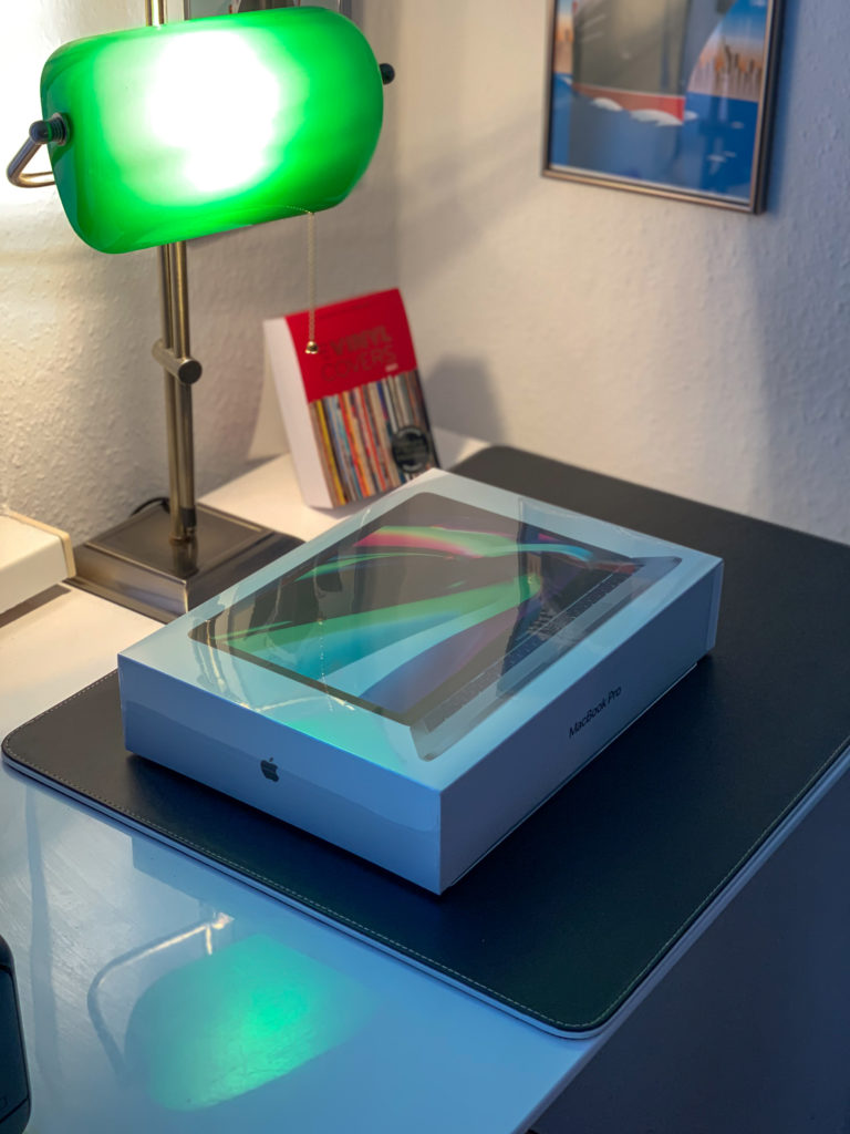 Ein MacBook liegt eingeschweißt in der Originalverpackung auf einem Schreibtisch, dahinter steht eine grüne Buchmacherlampe.