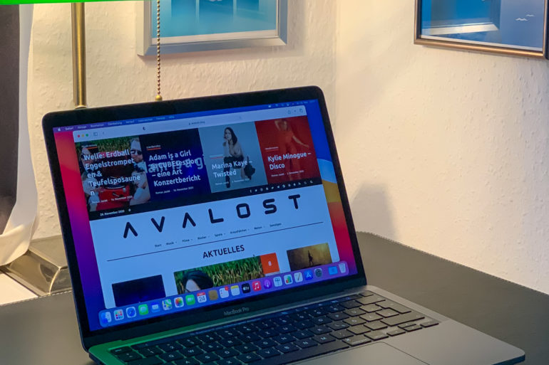 Ein graues MacBook Pro in 13 Zoll Größe steht aufgeklappt auf einem Schreibtisch. Der Webbrowser Safari ist geöffnet und zeigt die Webseite avalost.de an. Hinter dem MacBook steht eine grüne Buchmacherlampe auf dem Schreibtisch.