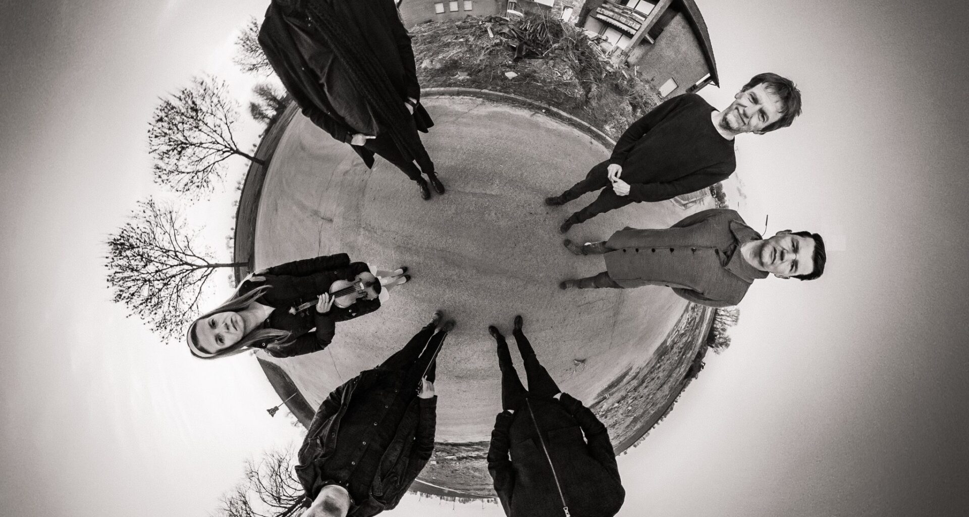 Die Band NOYCE™ steht im Kreis auf der Straße eines verlassenes Dorfes. Das Bild ist von der Mitte aus aufgenommen und simuliert eine Art Fischaugenoptik.