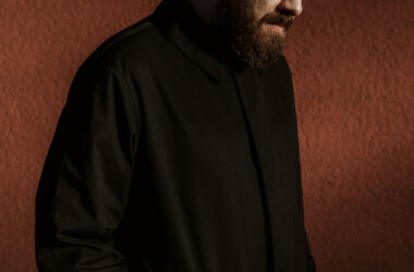 Ein Portrait von Ben Lukas Boysen. Er steht seitlich von der Kamera abgewandt, hat leicht lächelnd den Kopf gesenkt und trägt einen schwarzen Mantel.