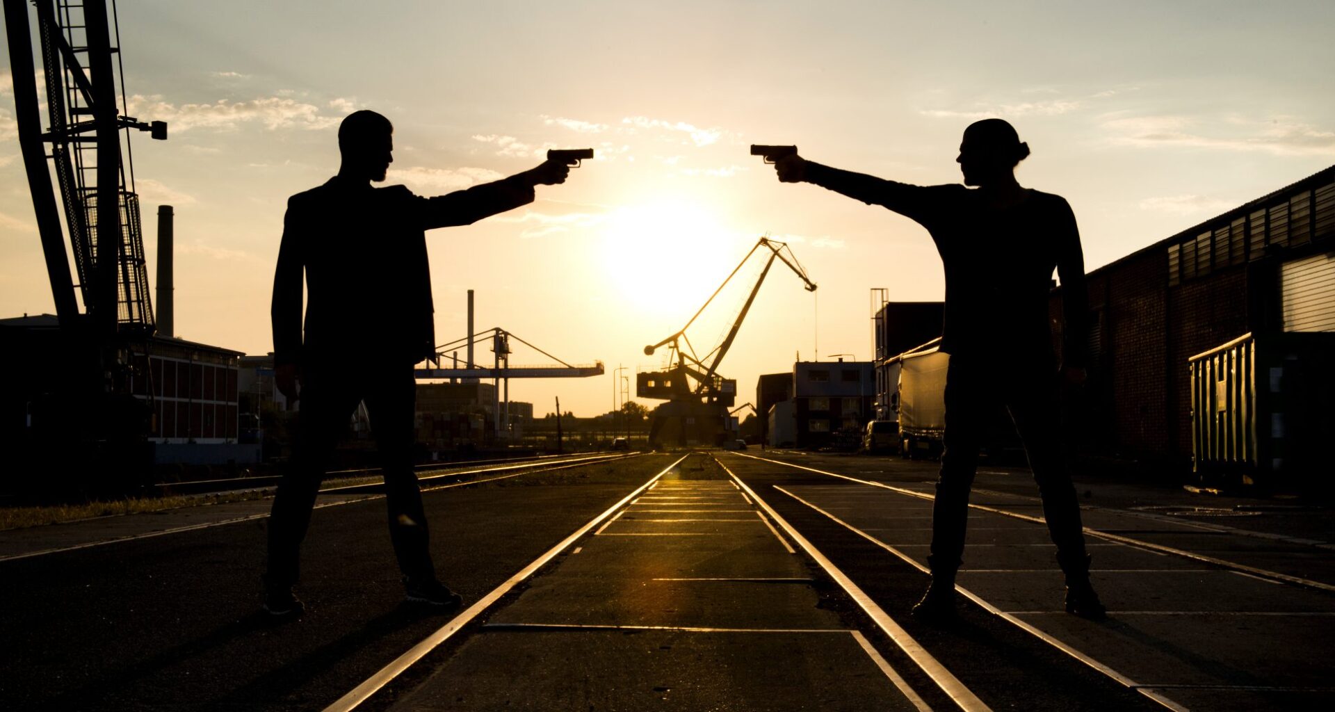 Ein Foto von Adrian Hates und Torben Wendt alias Coma Alliance. Sie befinden sich in einem Industriegebiet, im Hintergrund geht die Sonne langsam unter. Sie stehen sich gegenüber und bedrohen sich gegenseitig mit Pistolen, sind dabei aber lediglich als Schatten zu erkennen.