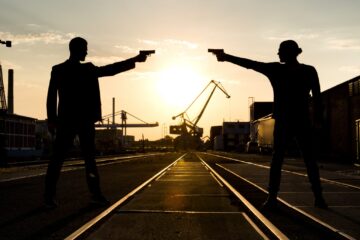 Ein Foto von Adrian Hates und Torben Wendt alias Coma Alliance. Sie befinden sich in einem Industriegebiet, im Hintergrund geht die Sonne langsam unter. Sie stehen sich gegenüber und bedrohen sich gegenseitig mit Pistolen, sind dabei aber lediglich als Schatten zu erkennen.