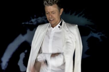 David Bowie vor dunklem Hintergrund, er trägt ein weißes Hemd und ein locker über die Schultern gelegtes Jackett.