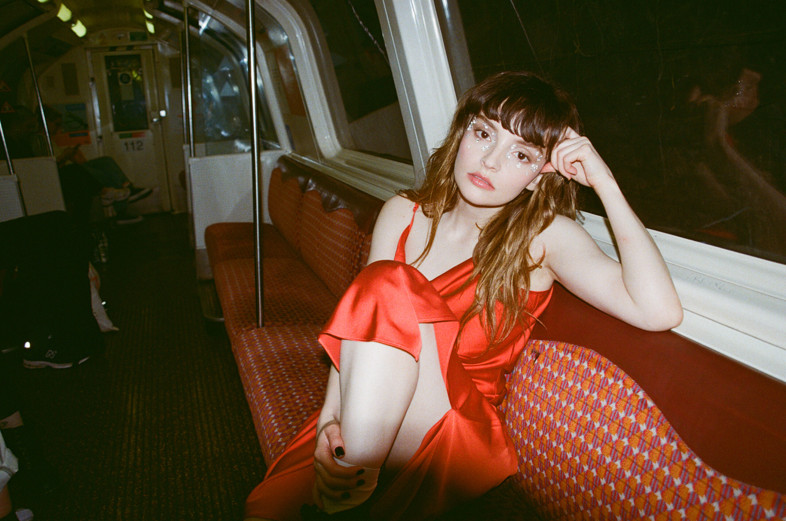 Lauren Mayberry sitzt bequem in einer U-Bahn, sie trägt ein rotes, glänzendes Kleid.